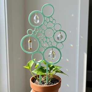 May include: A mint green plant trellis featuring a pattern of circles and paw prints, with three hanging clear crystal prisms and one small gold-toned cylindrical vial suspended from the circular frames, inserted into a terracotta plant pot.