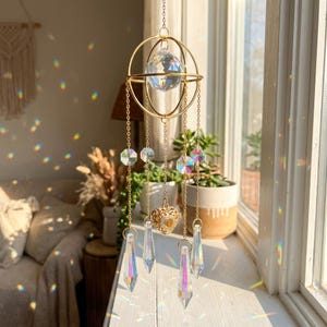 May include: A gold-colored hanging crystal suncatcher with a large crystal ball at the top, surrounded by a gold frame. Several strands of crystals and a heart-shaped charm hang below. The crystals are clear and reflect rainbow light.