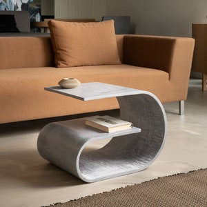 May include: A light grey concrete side table with a curved design and two shelves. The table is in a living room setting next to a brown sofa.