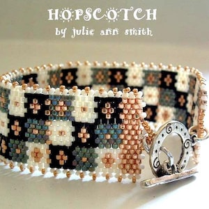 May include: A beaded bracelet with a black, white, and copper color scheme. The bracelet has a toggle clasp and is labeled "HOPSCOTCH by julie ann smith".