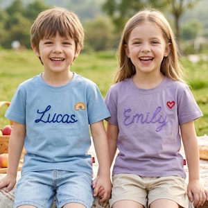 Puede incluir: Una camiseta azul claro con el nombre Lucas bordado en hilo azul oscuro y un pequeño parche de arcoíris, y una camiseta morado claro con el nombre Emily bordado en hilo morado y un pequeño parche de corazón rojo, ambas de manga corta.