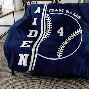 May include: A navy blue fleece blanket with a white baseball design. The baseball has white stitching and the number "4" in the center. The text "AIDEN" is on the left side of the blanket and "TEAM NAME" is on the right side.