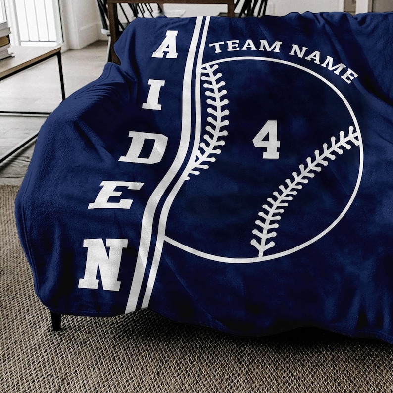 May include: A navy blue fleece blanket with a white baseball design. The baseball has white stitching and the number "4" in the center. The text "AIDEN" is on the left side of the blanket and "TEAM NAME" is on the right side.