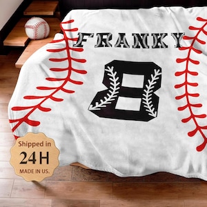 Puede incluir: Manta blanca con temática de béisbol con costuras rojas y el nombre "FRANKY" en negro. Un gran número negro "8" está centrado, también con detalles blancos. Una pelota de béisbol descansa sobre una superficie de madera.