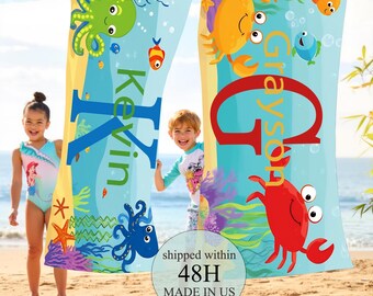May include: Two personalized beach towels with ocean-themed designs. One towel features a large blue letter "K" with the name "Kevin" and an octopus. The other towel has a red letter "G" with the name "Grayson" and a crab. The towels are hanging on a clothesline.