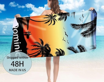 May include: Beach towel with a vibrant sunset design featuring palm trees and silhouettes. The towel transitions from orange to yellow to blue, with the word "Dominic" printed vertically. The towel is held by a person on a beach.
