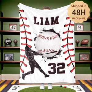 May include: White fleece blanket with a baseball theme. A baseball player is depicted swinging a bat, breaking through a wall with a baseball behind him. The blanket has red stitching around the edges, a baseball graphic, and the name "LIAM" and number "32" printed on it.