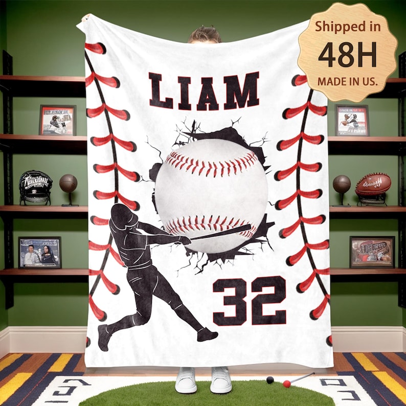 May include: White fleece blanket with a baseball theme. A baseball player is depicted swinging a bat, breaking through a wall with a baseball behind him. The blanket has red stitching around the edges, a baseball graphic, and the name "LIAM" and number "32" printed on it.