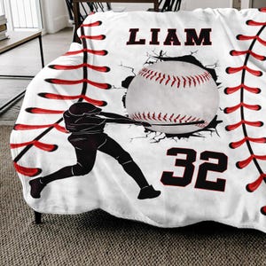 May include: White baseball themed fleece blanket with red stitching, a baseball breaking through the stitching, a baseball player silhouette, the name "LIAM" and the number "32" in black.