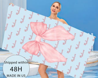 May include: A light blue beach towel with a large pink bow design. The towel is patterned with the name "Madison" in a repeating script. The towel is held up against a blue background.