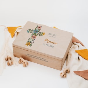 Peut inclure: Un coffret en bois clair orné d'une croix florale aux couleurs pastel. Le coffret porte les inscriptions "DEIN TEXT", "Maura" et "01. MAI 2025". Des jouets en bois et du tissu décorent la scène.