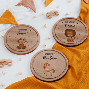 Puede incluir: Tres platos redondos de madera con nombres grabados e ilustraciones de animales: un mono, un león y un unicornio. Cada plato tiene el texto "Hier Krümelt" encima del nombre, con lunares de colores.