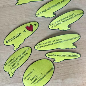 Könnte beinhalten: Eine Sammlung von hellgrünen Aufklebern in Sprechblasenform mit schwarzem Text. Die Aufkleber zeigen programmbezogene Phrasen wie "#include <love>", "works on my machine" und "emotional damage". Einige Blasen haben ein rotes Herz.