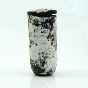 May include: A cylindrical, handmade ceramic container with a dark, textured lid. The body is primarily white with dark, mottled markings and a rough, aged appearance. The container has a slightly rounded base.