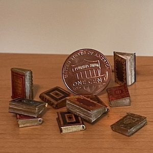 Può includere: Una collezione di libri in miniatura di vari colori e design, tra cui copertine rosse, marroni e nere con dettagli dorati, disposti attorno a una moneta statunitense da un centesimo per scala, con il testo United States of America.