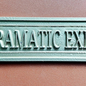 May include: A rectangular metal sign with a silver finish and a beaded border, featuring the raised text "DRAMATIC EXIT" in capital letters. The sign is secured with two visible screws on the left and right sides against a solid brown surface.