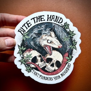 Puede incluir: Una pegatina con una ilustración de una zarigüeya con la boca abierta, mostrando los dientes, y dos calaveras. El texto "BITE THE HAND THAT MURDERS YOUR NEIGHBORS" está en un borde circular alrededor de la imagen. La paleta de colores incluye gris, blanco y rojo.