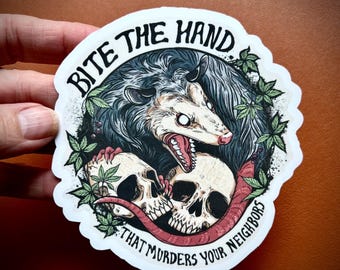 Puede incluir: Una pegatina con una ilustración de una zarigüeya con la boca abierta, mostrando los dientes, y dos calaveras. El texto "BITE THE HAND THAT MURDERS YOUR NEIGHBORS" está en un borde circular alrededor de la imagen. La paleta de colores incluye gris, blanco y rojo.