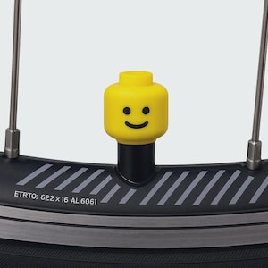 Könnte beinhalten: Eine gelbe Ventilkappe im Lego-Stil mit einem lächelnden Gesicht ist an einem Fahrradrad befestigt. Das Rad ist schwarz mit silbernen Akzenten und Speichen. Der Reifen trägt die Aufschrift "ETRTO: 622 x 16 AL 6061".