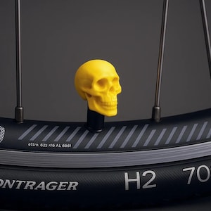 Puede incluir: Una tapa de válvula en forma de calavera amarilla en un neumático de bicicleta negro con el texto "BONTRAGER" y "H2 700x25" impreso en el lateral.