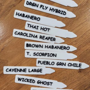 May include: White plant markers with black text listing different types of hot peppers. The names include: Hot Cherry, Drgn Fly Hybrid, Habanero, Thai Hot, Carolina Reaper, Brown Habanero, T. Scorpion, Pueblo Grn Chile, Cayenne Large, Wicked Ghost, and Mucho Jalapeno.