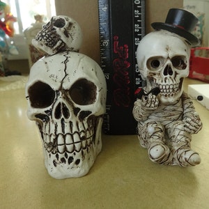 May include: Two white skull figurines. One skull is wearing a black top hat and is sitting down. The other skull is standing up and has a smaller skull on top of its head.