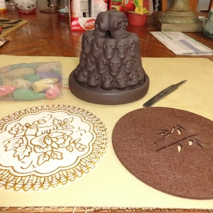 May include: A brown ceramic incense burner shaped like a stack of skulls. The burner is on a table with a small bag of incense cones, two coasters, and a small metal tool.