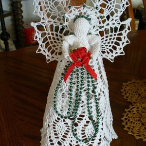 May include: A handmade white crochet angel figurine with detailed openwork wings and a flowing gown. The angel holds a bouquet of red roses and is adorned with green beaded accents and a red ribbon. A decorative item.
