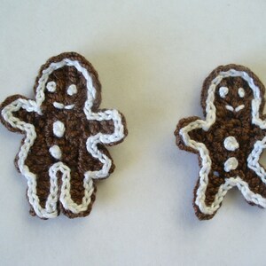 May include: Two brown crocheted gingerbread people with white icing details. The gingerbread people are standing on a white surface.