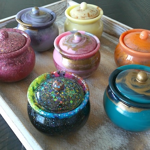 May include: Seven small decorative jars with lids. The jars are made of resin and have a variety of colours and patterns, including pink, teal, black, orange, yellow, and purple. Each jar has a gold accent on the lid.