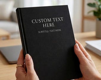 May include: A black hardcover notebook with white text printed on the front cover that reads CUSTOM TEXT HERE and SUBTITLE TEXT HERE. The notebook is held upright, displaying its solid black matte finish and clean, professional design.