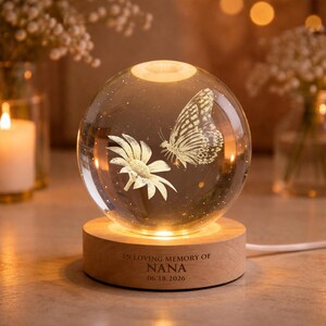 Puede incluir: Una esfera de vidrio transparente con un diseño de mariposa y flor, sobre una base de madera. La base tiene la inscripción "IN LOVING MEMORY OF NANA 06.18.2026". La esfera está iluminada desde dentro, creando un brillo cálido.