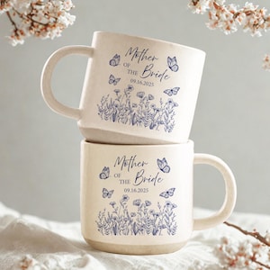 Könnte beinhalten: Zwei cremefarbene Keramikbecher, die übereinander gestapelt sind. Jeder Becher hat ein blaues Blumen- und Schmetterlingsdesign mit dem Text "Mother of the Bride 09.16.2025". Die Becher haben ein rustikales, strukturiertes Aussehen.