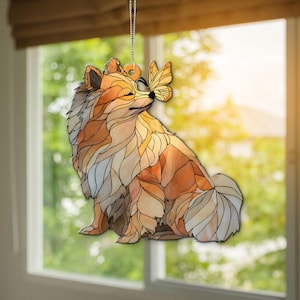 Può includere: Ornamento decorativo sospeso raffigurante un cane Pomerania con una farfalla. Il cane è raffigurato in tonalità arancioni, marroni e crema, con una farfalla appoggiata sul naso. L'ornamento è appeso a una catena d'argento.