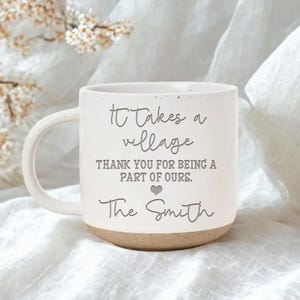 Puede incluir: Una taza de cerámica blanca con acabado moteado y base color canela. El texto dice: It Takes a village, THANK YOU FOR BEING A PART OF OURS, un pequeño icono de corazón y The Smith. La taza presenta una mezcla de letras cursivas y de molde.