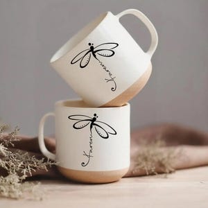 Op de afbeelding: Twee witte keramische mokken met een bruine basis, elk met een zwart libellenontwerp en de naam "Karen" in script. De mokken hebben een eenvoudig, elegant ontwerp met een gebogen handvat.