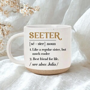 Könnte beinhalten: Weiße Keramik-Tasse mit dem Wort "SEETER" in goldfarbenen Buchstaben. Auf der Tasse steht eine Definition: "Wie eine normale Schwester, aber viel cooler" und "Beste Freundin fürs Leben."