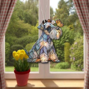 May include: A decorative hanging ornament featuring a Schnauzer dog design with a butterfly. The dog is rendered in a stained-glass style, with blue, purple, and orange hues. It hangs from a silver chain, perfect for a window or rearview mirror.