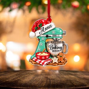 May include: A decorative ornament shaped like a mint green stand mixer wearing a red Santa hat with the name Anna. The mixer features a silver bowl labeled Baking Queen, a red bowl with a wooden spoon, a red plaid cloth, and a gingerbread cookie.