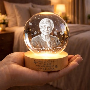 Könnte beinhalten: Personalisierte Kristallkugel mit einem Porträt einer Frau, dem Wort "Mom" und den Jahren 1956-2026. Die Kugel steht auf einem Holzsockel mit der Inschrift "Die, die wir lieben, gehen nicht weg, sie gehen jeden Tag an unserer Seite."