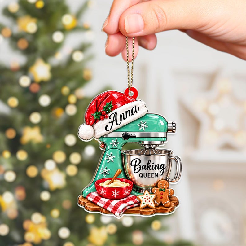 May include: A flat Christmas tree ornament shaped like a teal stand mixer with a red Santa hat on top. The mixer features the name Anna, a silver bowl labeled Baking Queen, a red bowl with dough, a red plaid cloth, and a gingerbread cookie.