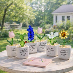 Könnte beinhalten: Eine Sammlung von Glasblumenskulpturen in kleinen weißen Töpfen, darunter eine rosa Tulpe, eine blaue Hyazinthe und eine gelbe Sonnenblume. Jeder Topf trägt den Text "Never forget the difference you make" und ein Herz. Eine rosa Karte liegt auf dem Tisch.
