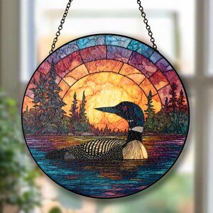 May include: Round stained glass panel featuring a loon on a lake at sunset. The artwork has a colorful mosaic design with a black chain for hanging. The loon is black, white, and tan, with a red eye.