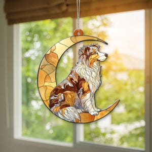 Könnte beinhalten: Ein dekorativer Fensterhänger mit einem Hund, der auf einem Halbmond sitzt. Der Hund hat braunes, weißes und hellbraunes Fell, und der Mond ist ein Mosaik aus warmen Farben. Der Fensterhänger hängt an einer silbernen Kette.