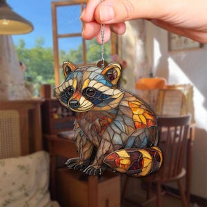 May include: A colorful raccoon ornament with a stained glass effect. The raccoon is depicted in shades of brown, orange, blue, and yellow. It is hanging from a silver chain. The ornament is a decorative item.