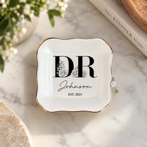 May include: A square white ceramic trinket dish with scalloped edges and a gold-painted rim. The centre features black text reading DR, Johnson, and EST. 2025, with floral line art integrated into the letters. A silver ring rests on the right edge.