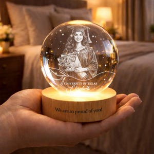 Könnte beinhalten: Eine klare Glaskugel mit einem Abschlussporträt, Sternen und dem Text "University of Texas Elizabeth" steht auf einem Holzsockel. Der Sockel ist mit "We are so proud of you!" graviert. Die Kugel wird warm beleuchtet.