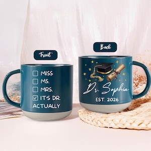 Könnte beinhalten: Türkisfarbene Keramiktasse mit grauem Boden. Auf der Vorderseite steht "MISS, MS., MRS., IT'S DR. ACTUALLY" mit einem Häkchen. Die Rückseite zeigt eine Doktorhut, eine Schriftrolle, Sterne und "Dr. Sophia EST. 2026".