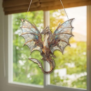 May include: A stained glass dragon suncatcher with outstretched wings, suspended by a chain. The dragon is detailed with brown, gold, and blue hues. The wings are a mosaic of light-coloured glass, creating a beautiful display. The dragon is hanging in front of a window.