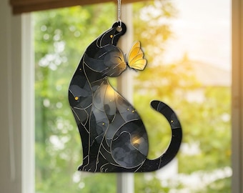 Puede incluir: Adorno en forma de gato negro con una mariposa amarilla. El gato está sentado, con una mariposa posada en su nariz. El cuerpo del gato tiene un efecto de vidriera con detalles dorados. El adorno cuelga de una cadena plateada.